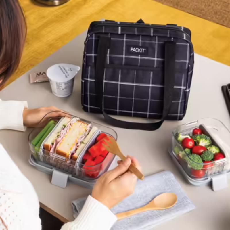 PackIt freezable lunch bag in Hampton style in Black Grid design - on desk with woman eating food. 